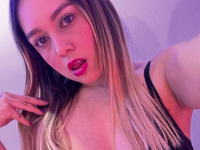 AmayaJhons has pinkish skin, red lips, and blonde hair, wearing a black top, with her hand near her mouth, looking into the camera. Her tits are partially visible.