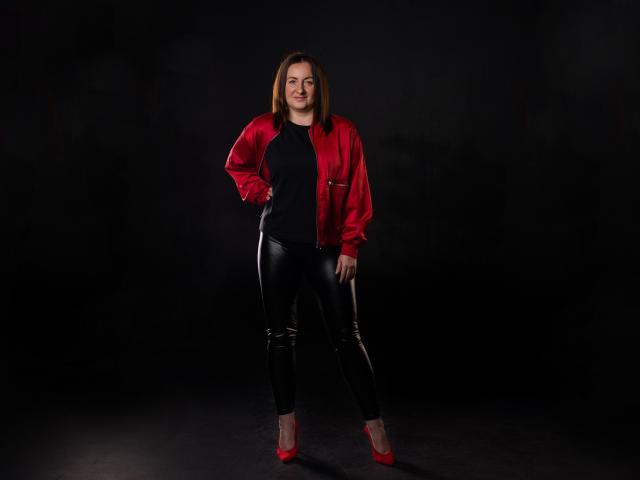 AbbyTaylorr stands in a dark room, wearing a red satin jacket, black leather pants, and red heels, with hands on hips, smiling slightly, and looking confident.