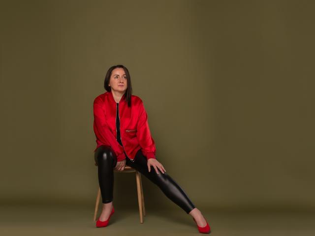 AbbyTaylorr is sitting on a wooden chair, wearing a red satin jacket, black shiny leggings, and red heels, looking up with a serious expression.