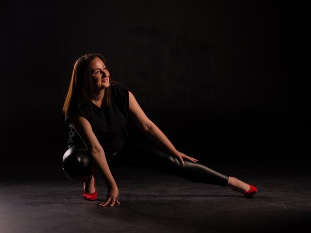 AbbyTaylorr, squatting with black leather pants, red heels, and black top, stretching her right leg, smiling, brown hair, dark background, looking happy and relaxed.