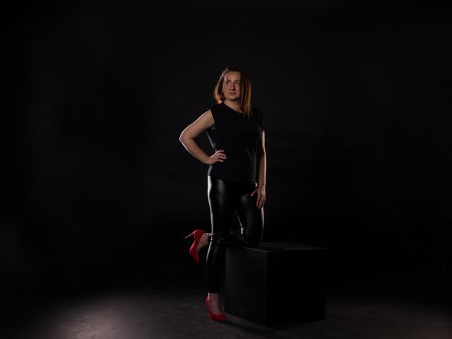 AbbyTaylorr stands with one knee on a black box, wearing a black top, shiny black leather pants, and red high heels. She has a hand on her hip.