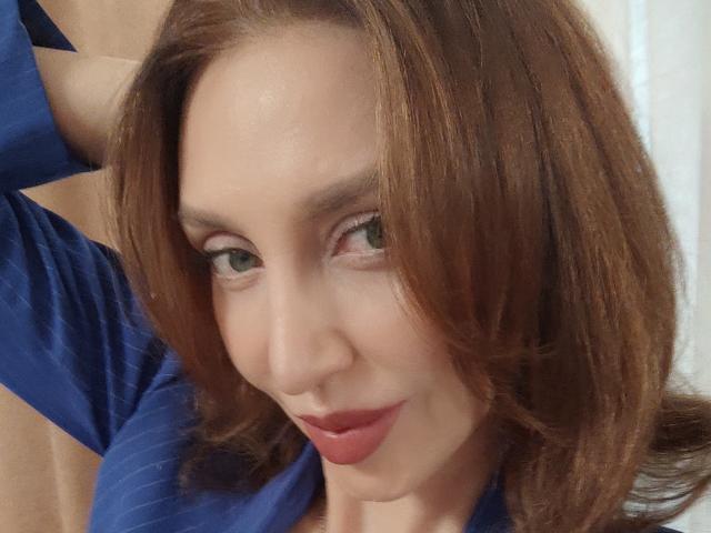 SimoniGrace, con i capelli castani, gli occhi verdi e il rossetto rosso, indossa una maglietta blu, ha un braccio alzato, sorride dolcemente, mostrando il suo viso carino e il suo piccolo naso..