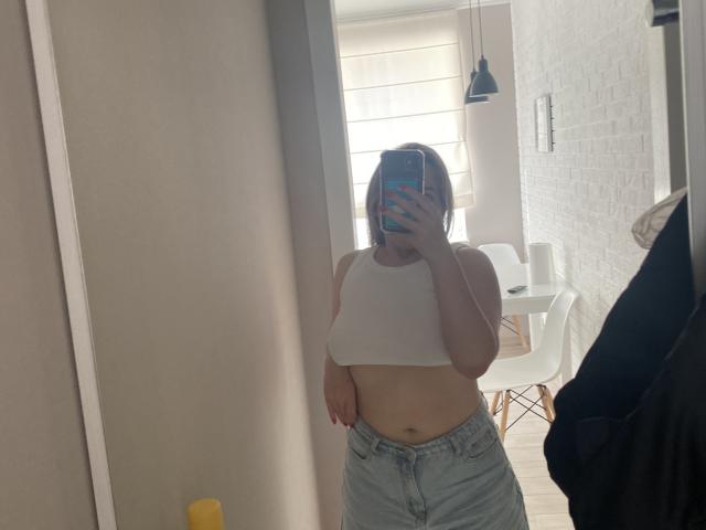 EmberSexy, wearing a white crop top and high-waisted jeans, shows her midriff in a mirror selfie. Her hand covers her phone, showing only her face.