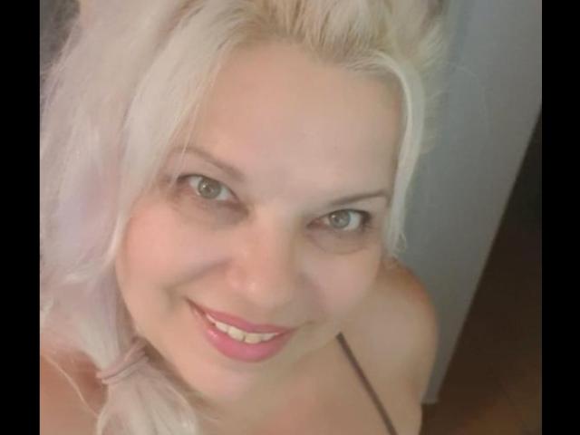 SoniaHotMilf with platinum blonde hair, smiling at the camera, showing pink lipstick and cute green eyes, wearing a black strap, looking down. Her boobs are partially visible.