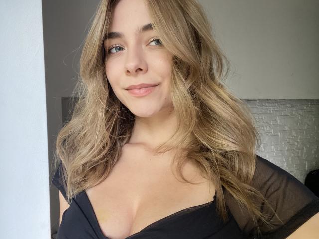 AmyTasty tiene el pelo ondulado de color castaño claro, ojos azules y una sonrisa discreta. Lleva una blusa negra transparente que revela su escote y sus pequeños senos. Está de pie en una habitación bien iluminada..