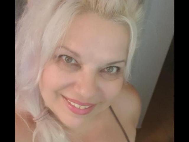 SoniaHotMilf with blonde hair, pink lipstick, smiling at the camera, looking up, showing her tits, with green eyes, fair skin, and a cute face.