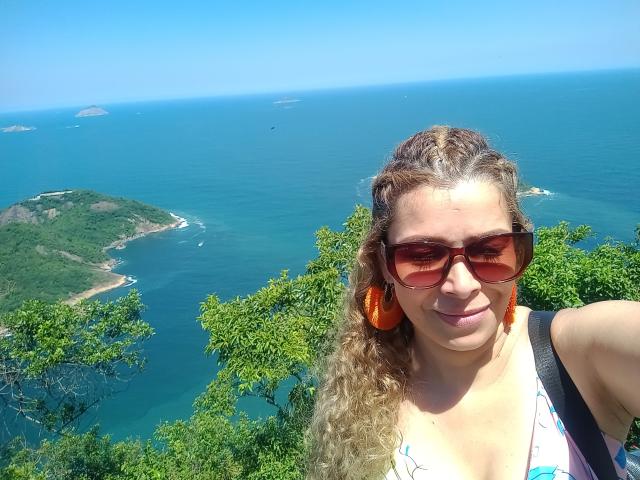 Dalila, muito atraente, com cabelos loiros cacheados, óculos de sol vermelhos grandes, brincos laranja e uma blusa azul, mostrando os seios, sorrindo, em pé perto de uma árvore, com um oceano azul ao fundo..