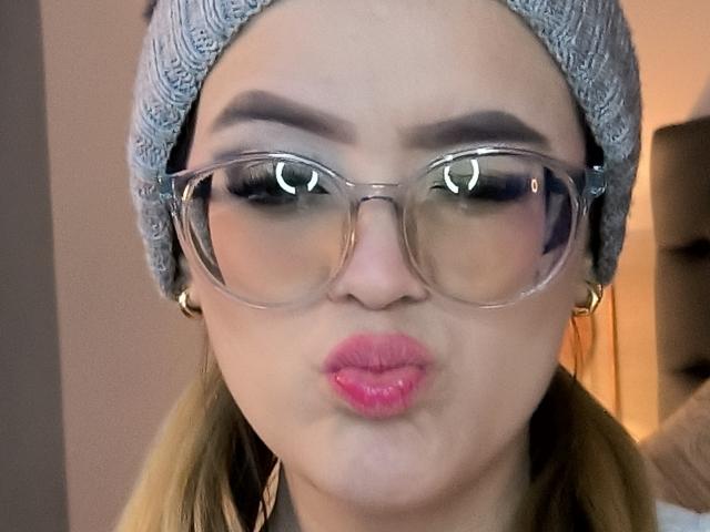 Kim, en été, avec des lèvres roses et gonflées, des lunettes transparentes et un bonnet en tricot gris, faisant une moue. Elle a les cheveux blonds, des sourcils foncés et des boucles d'oreilles dorées..