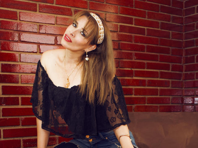 DalilaSuperHot with long brown hair, wearing a black lace off-shoulder top, blue jeans, and a patterned headband, red lipstick, gold necklace, and earrings, leaning against a red brick wall.