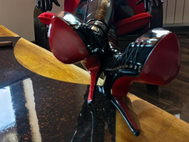 GoddessKaterina's shiny black latex boots with red soles are prominently displayed in the foreground, her legs crossed, showing the bottom of her high heels.