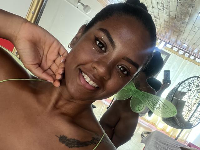 TiffanySammy with dark skin, smiling, green butterfly earrings, black tattoo on right shoulder, brown eyes, black hair in bun, wearing green strap, holding right cheek.