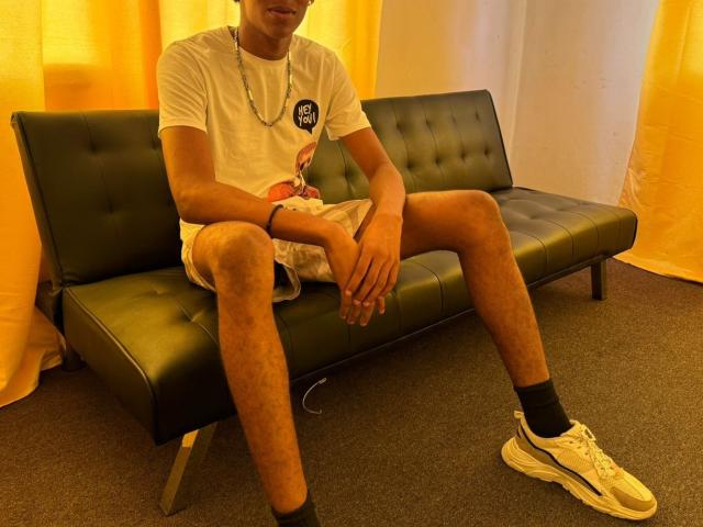 Hakeem sitzt auf einem grünen, gepolsterten Sofa, trägt ein weißes T-Shirt, beige Shorts, schwarze Socken, weiße Turnschuhe und eine blaue Halskette, mit gespreizten Beinen und ruhenden Armen..