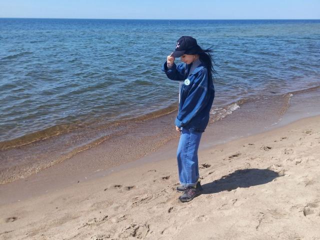Zanoza steht an einem sandigen Strand, trägt eine blaue Jacke, Jeans, eine schwarze Kappe und schwarze Schuhe, und ihr langes, schwarzes Haar weht im Wind, in der Nähe sanfter Wellen..
