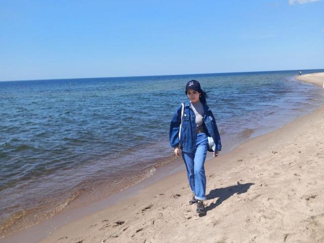 Zanoza cammina su una spiaggia sabbiosa, indossando una giacca blu, jeans e scarpe nere, con un cappello blu e una collana bianca, sullo sfondo di un cielo e un mare azzurri..