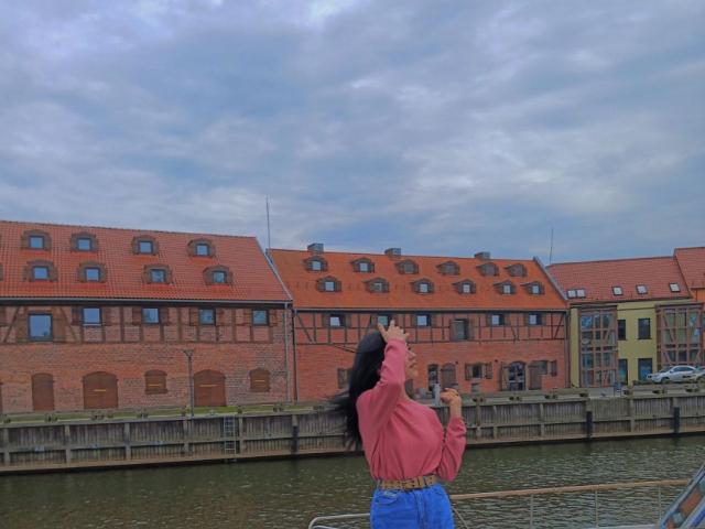 Zanoza, con lunghi capelli neri, una camicia a maniche lunghe rossa e pantaloni blu, in piedi vicino a un fiume, con una sigaretta in mano, di fronte a edifici in mattoni con tetti rossi..
