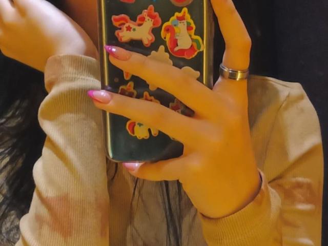 La mano de Zanoza, con uñas rosas, sosteniendo un teléfono con pegatinas de unicornios, lleva un anillo y un suéter beige, visible desde el cuello hacia abajo, que cubre ligeramente su rostro..
