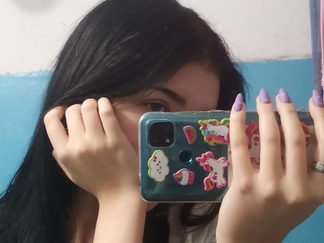 Zanoza with long black hair, purple nails, holding a phone with colorful stickers, partially covering her face, taking a mirror selfie. Her eye is visible.