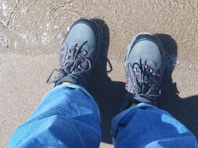 Zanoza's achterkant, in blauwe broek, met grijze en roze veterschoenen, staat op nat zand met kleine waterdruppels..