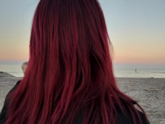 Zanoza heeft rood, recht haar van gemiddelde lengte, en je ziet haar van achteren. Ze staat op een strand met een zachte zonsondergang en een rustige oceaan op de achtergrond. Ze draagt een donker jasje..