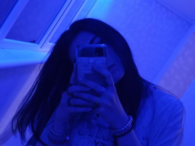 Zanoza, com cabelo longo e escuro, selfie no espelho com tons de azul, segurando o celular cobrindo o rosto, vestindo uma camisa branca, pulseiras, um anel, luz azul, reflexo na janela..