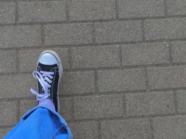 Zanoza's blauwe jeans en zwarte sneakers met witte accenten, licht beschadigd en met roze veters, staan op een ruwe, grijze bakstenen vloer..
