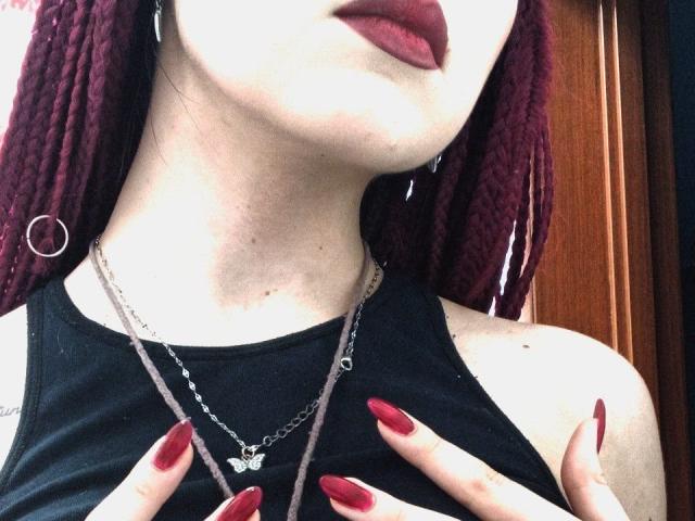 A Zanoza tem dreadlocks marrons, batom vermelho e unhas vermelhas, vestindo uma blusa preta com um colar de borboleta, segurando os seios. Ela tem brincos de argola prateados e um colar de corrente..