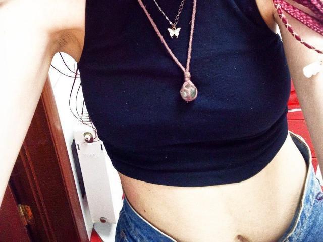 Zanozas schwarzes Crop-Top, das ihre Brüste zeigt, mit einer pinken Halskette und einem Kristallanhänger, und blaue Jeans, die Arme leicht angehoben. Ihre Achselhöhlen sind sichtbar..