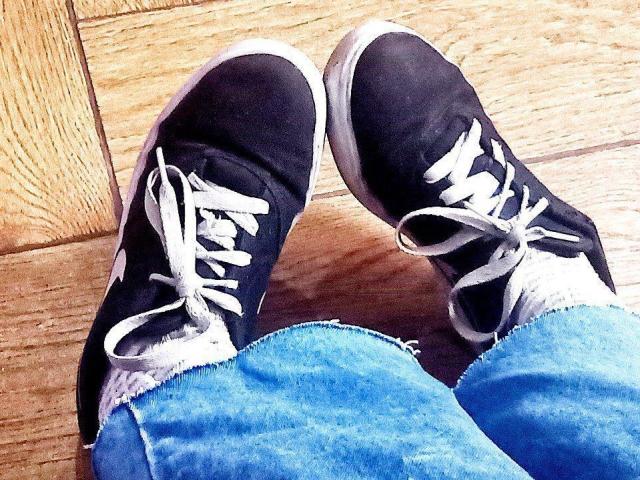 Le scarpe da ginnastica nere di Zanoza, con lacci bianchi e bordi sfilacciati, poggiate su un pavimento di legno, indossa jeans blu, leggermente strappati..