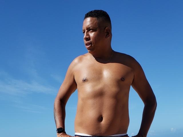 Douglas69 is a shirtless, muscular, dark-skinned man with a short, buzzed haircut, wearing white underwear and a black watch, standing against a clear blue sky.