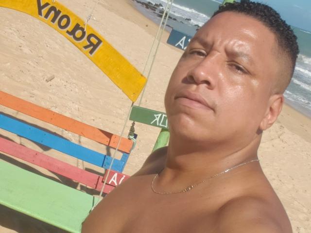 Douglas69, mit ernstem Gesicht, ohne Hemd, mit kurzen, lockigen Haaren, gebräunter Haut, einer kleinen Halskette, sitzend auf einer bunten Bank, Strand im Hintergrund, sonnig, leicht verschwitzt, blickt in die Kamera..