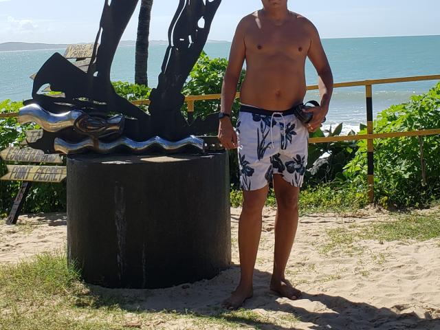 Douglas69 staat zonder shirt, gekleed in bloemenprint zwemshorts in wit en zwart, met een zwarte metalen sculptuur en een gele reling op de achtergrond, en een blauw-groene oceaan achter hem..