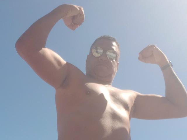 Douglas69, zonder shirt, met beide biceps gespannen, draagt reflecterende zonnebril, heeft een donkere huidskleur, kort haar en een gespierd lichaam, tegen een helderblauwe lucht..