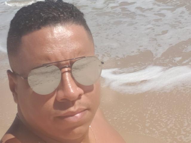 Douglas69 with short black hair, wearing reflective sunglasses, glistening skin, and a gold necklace, stands at the beach with foamy waves in the background.
