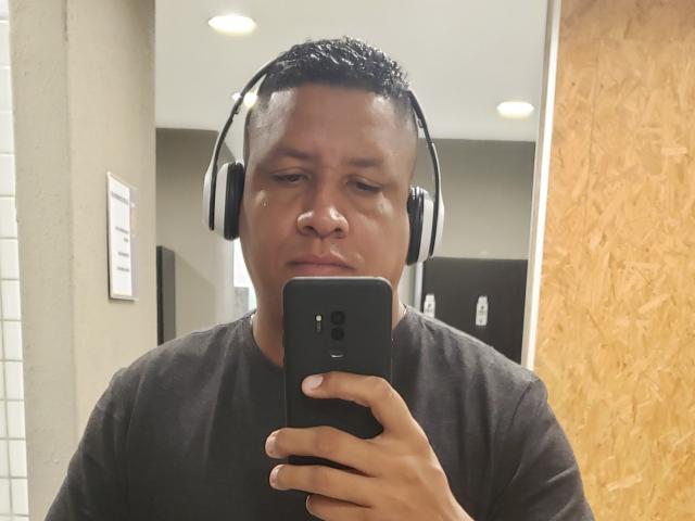 Douglas69, con i capelli neri corti, indossa una maglietta nera e delle cuffie, e sta scattando un selfie con un telefono nero in uno specchio. Il suo viso mostra un leggero accenno di smorfia..