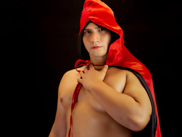 KingaNDher est un homme torse nu portant une capuche rouge en satin, agrippant l'avant, ce qui révèle sa poitrine nue et ses petits mamelons sur un fond sombre. Il a l'air sérieux..