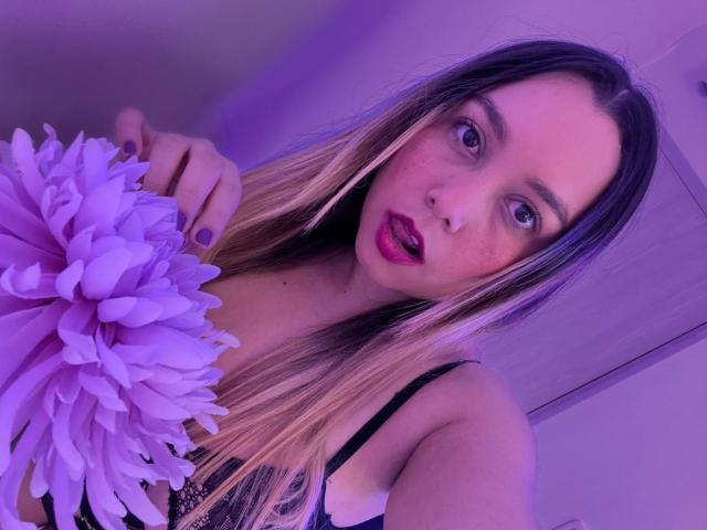 AmayaJhons holding a purple flower, with long blonde hair, purple lipstick, and a black lace bra, looking into the camera with a slightly open mouth.