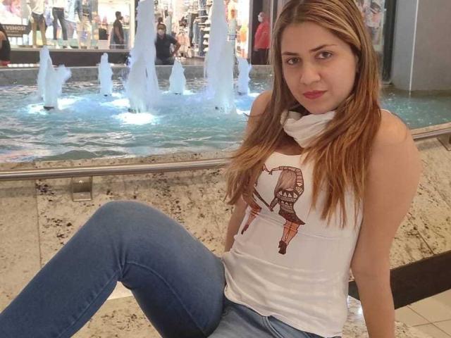 StevenAndPaty tem cabelos castanhos longos, uma camiseta branca com uma estampa marrom, e jeans azuis, sentada com as pernas cruzadas, mostrando os seios, em um shopping..
