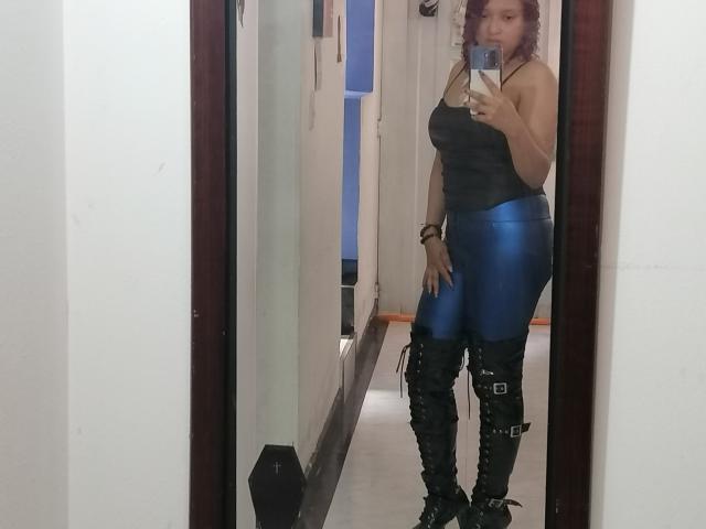 VioletaSwitch with purple hair, black tank top, and blue shiny leggings, wearing black lace-up boots with straps, taking a mirror selfie, standing in a hallway.