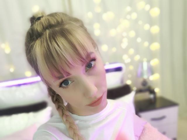 SweetyPretty with blonde braids, pink eyelids, and blue eyes, wearing a white shirt, looking up with a soft smile, in a softly lit bedroom with bokeh lights.