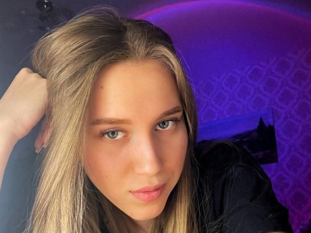 AmeliaAttom has blonde hair, blue eyes, and a slight pout. She's wearing a black top, with her hand in her hair, under purple lighting.