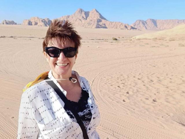 DoraBlueEyess with short brown hair, black sunglasses, white patterned shirt, black lace top, smiling, yellow backpack, desert with rocky mountains in the background, clear blue sky.