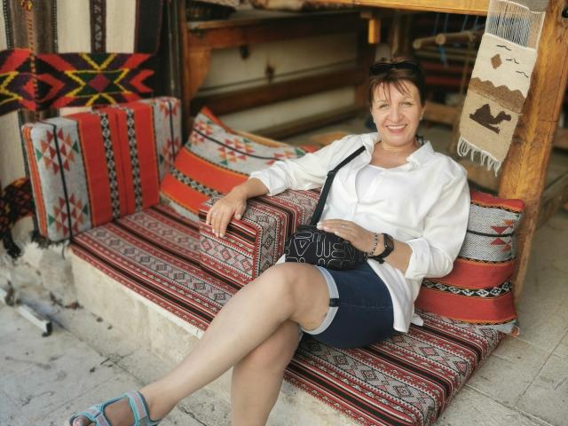 DoraBlueEyess with short brown hair, white shirt, blue shorts, black crossbody bag, and blue sandals, sitting on a red and white patterned bench, smiling. Her ass is partially visible.