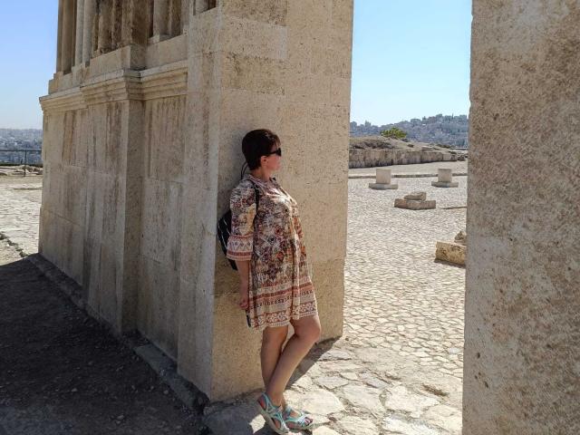 DoraBlueEyess stands against a stone pillar, wearing a patterned dress, sunglasses, and blue sandals, with a backpack, in a sunlit, ancient stone courtyard.