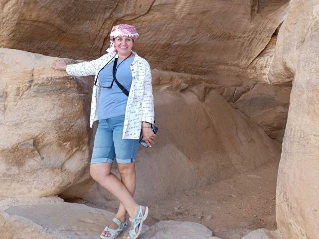 DoraBlueEyess stands in a rocky cave, wearing a pink bandana, white jacket, blue shirt, denim shorts, and sandals, with a camera strap across her chest.