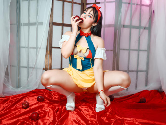 AngelCutie est assise sur du satin rouge avec des pommes, vêtue d'un costume bleu et jaune à une épaule, et suce une cerise rouge, exposant ses seins, avec des chaussettes blanches et des talons..