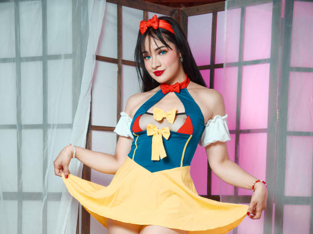AngelCutie com seios grandes, vestindo um vestido azul e amarelo, com um laço vermelho, mangas brancas, segurando a saia, batom vermelho, cabelo preto, tiara vermelha, fundo rosa e branco..