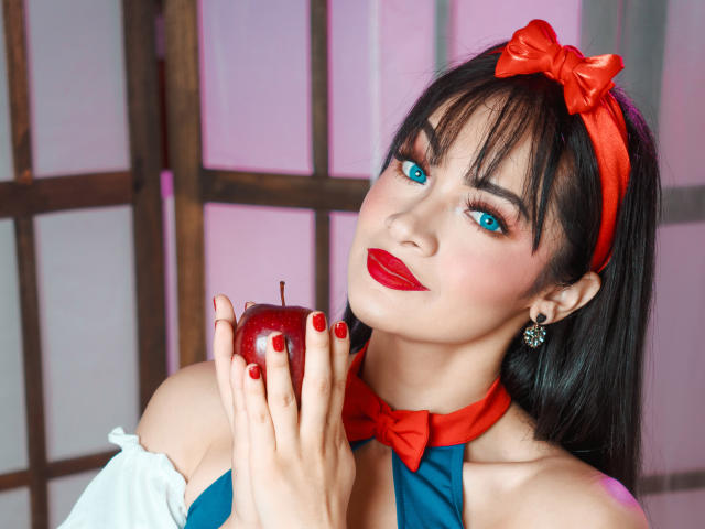 AngelCutie met rode lippenstift en blauwe ogen houdt een rode appel vast, draagt een rode strik in haar haar, een bijpassende choker en een blauw topje, waarbij haar borsten en rode nagellak te zien zijn..