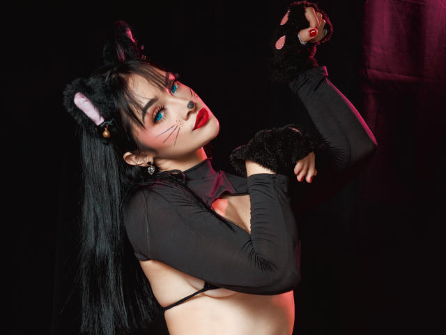 AngelCutie mit schwarzen Katzenohren, langen schwarzen Haaren und rotem Lippenstift, trägt ein schwarzes Oberteil mit langen Ärmeln, das die Brüste freilegt, sowie schwarze, pelzige Handschuhe, vor einem dunklen Hintergrund..