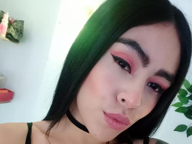 StellaBeckker, avec ses cheveux noirs, son fard à paupières rose, son collier noir, son piercing au nez et ses lèvres roses, regarde l'appareil photo, montrant son joli visage et son petit nez légèrement retroussé..