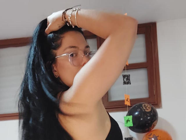 BiancaScott with black hair, glasses, and a black top, raising her arm to touch her hair, showing her tits and a bracelet on her wrist. Balloon in background.